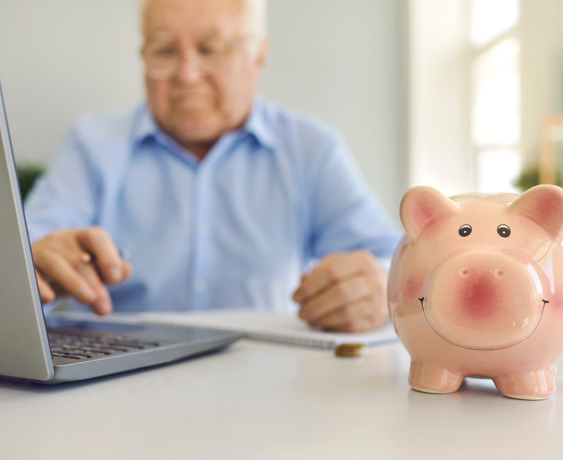 senior man on laptop computer in background with piggy bank in focus